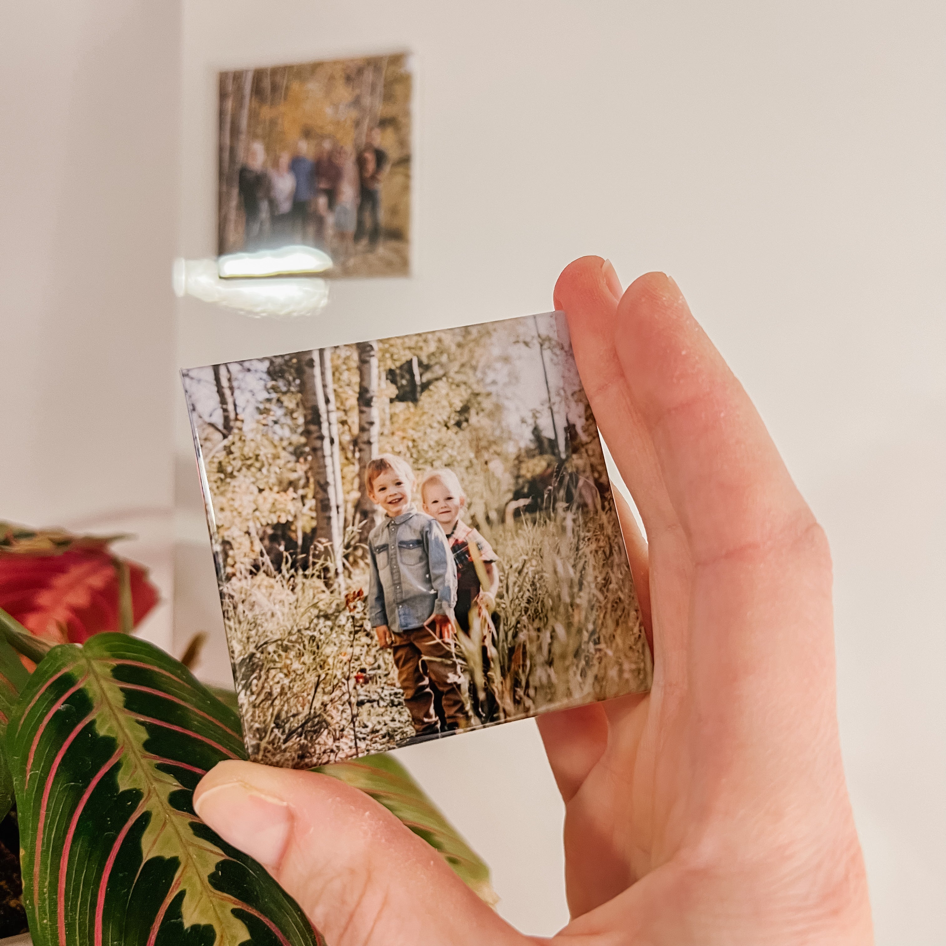 Hand holding a square custom photo fridge magnet of two children in nature, with a blurred indoor background.