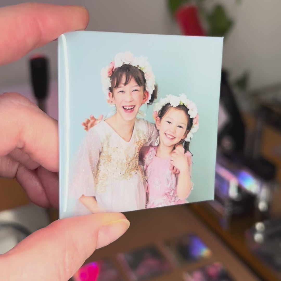Load video: A hand holding a square custom magnet showing 2 young girls smiling for the portrait.  There is a blurry background showing the magnet punch and press and some other custom magnets.